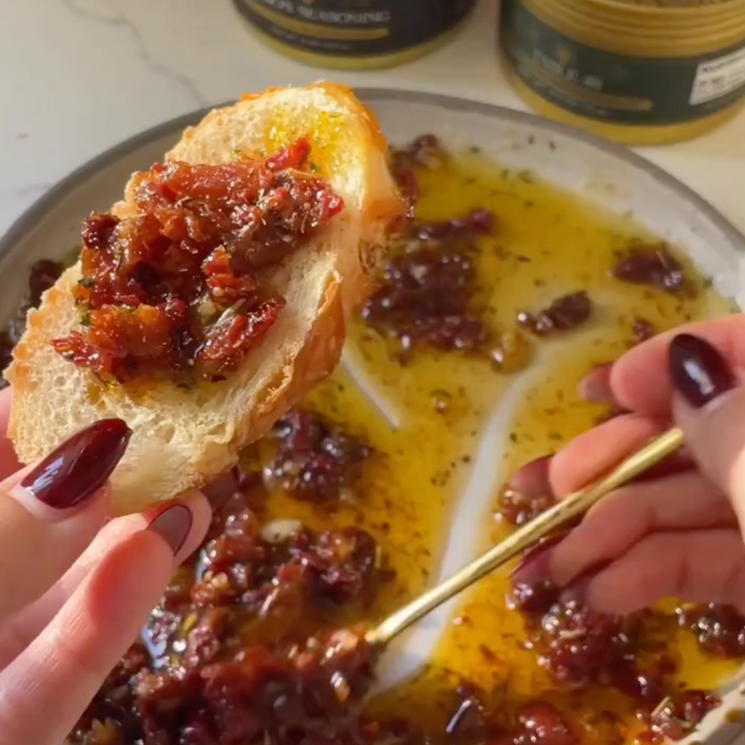 Hands tearing crusty bread to dip into sun-dried tomato Italian dipping oil with herbs.