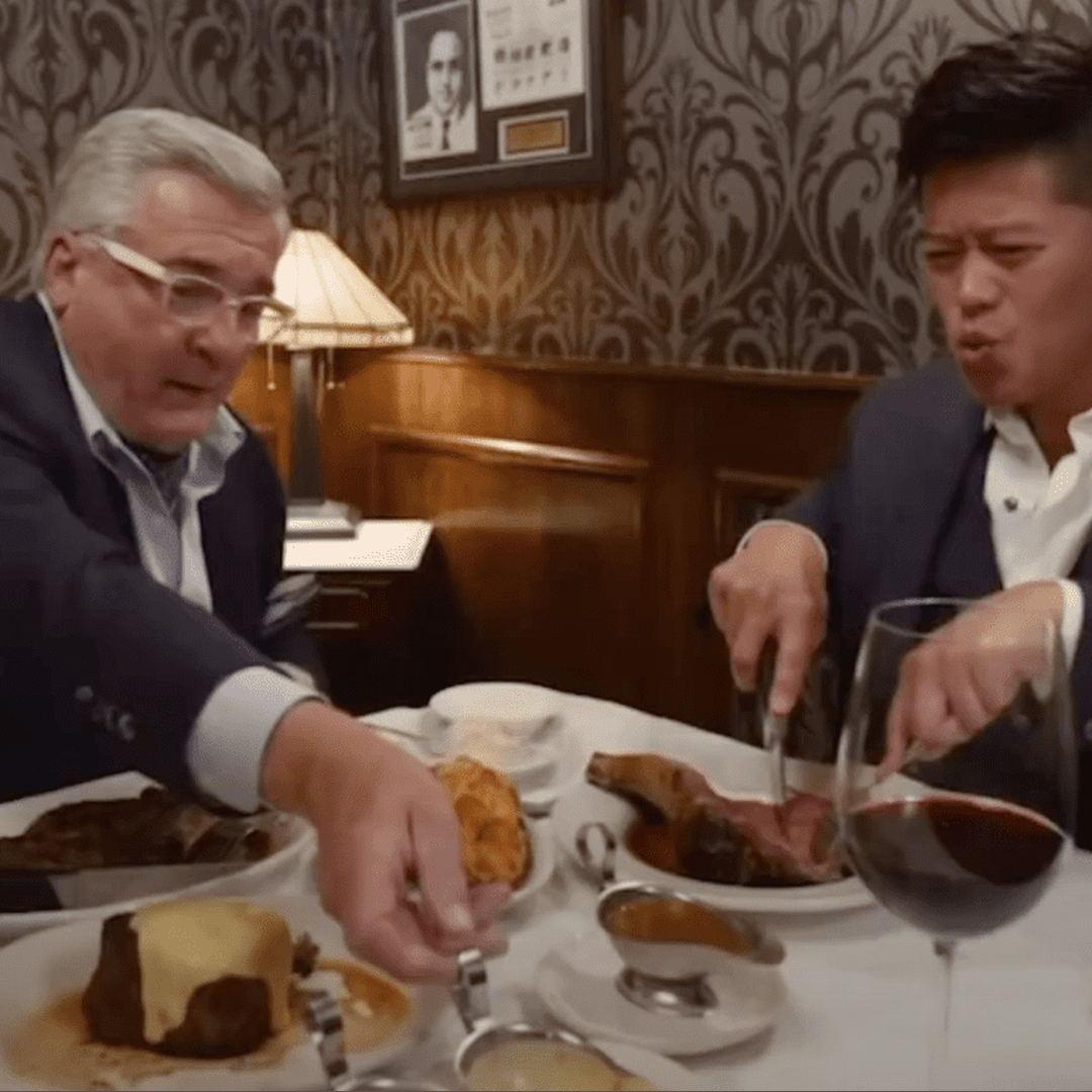 Two men dine at a wood paneled steakhouse table sharing plates of meat as one reaches for bread.