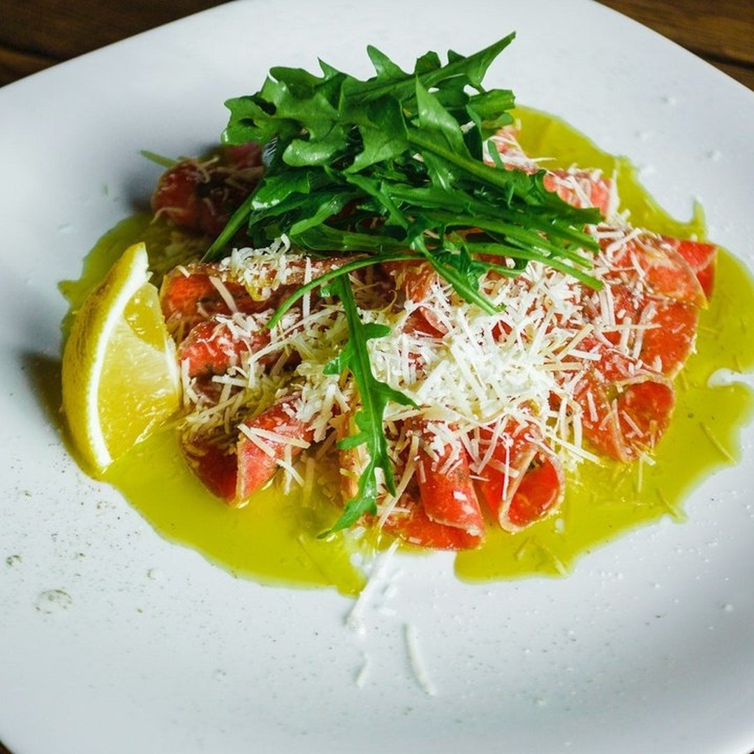 Beef carpaccio on a white plate topped with arugula and grated cheese with a lemon wedge on the side.