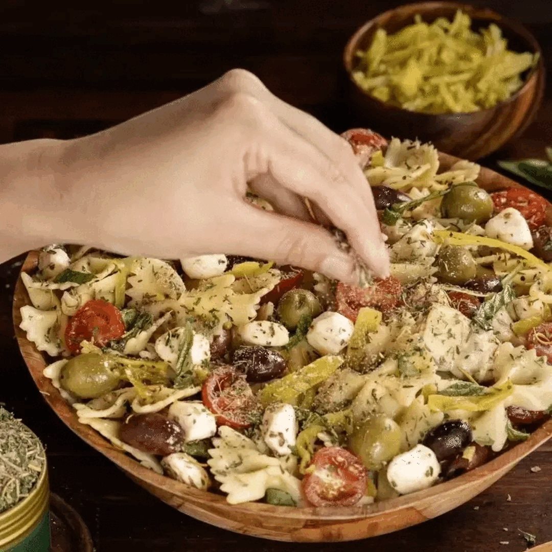 Hand sprinkling herbs over Mediterranean pasta salad with olives, cherry tomatoes and mozzarella in a wooden bowl.