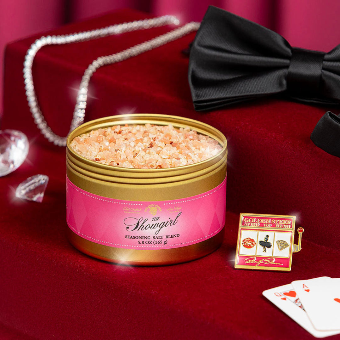 Pink The Showgirl seasoning blend container with gold lid on a red surface, surrounded by decorative items.