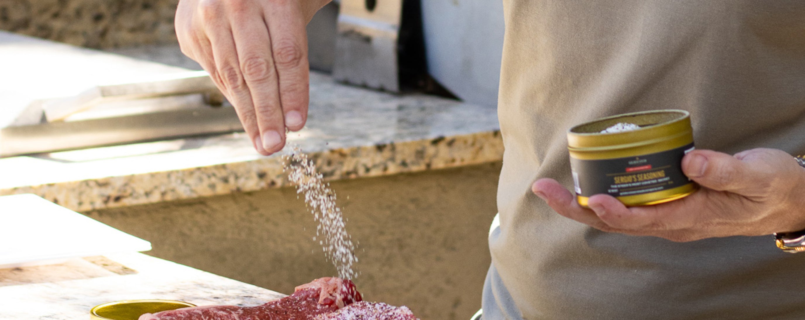 Hand sprinkling salt from a small tin labeled 'SERGIO'S SEASONING' onto raw steaks on a counter.