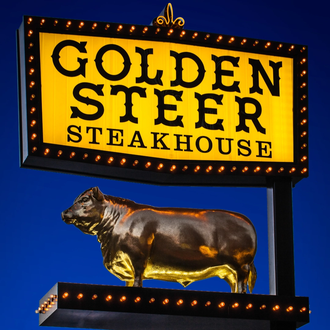 Illuminated Golden Steer Steakhouse sign reading 'GOLDEN STEER STEAKHOUSE' with a golden steer statue against a deep blue evening sky