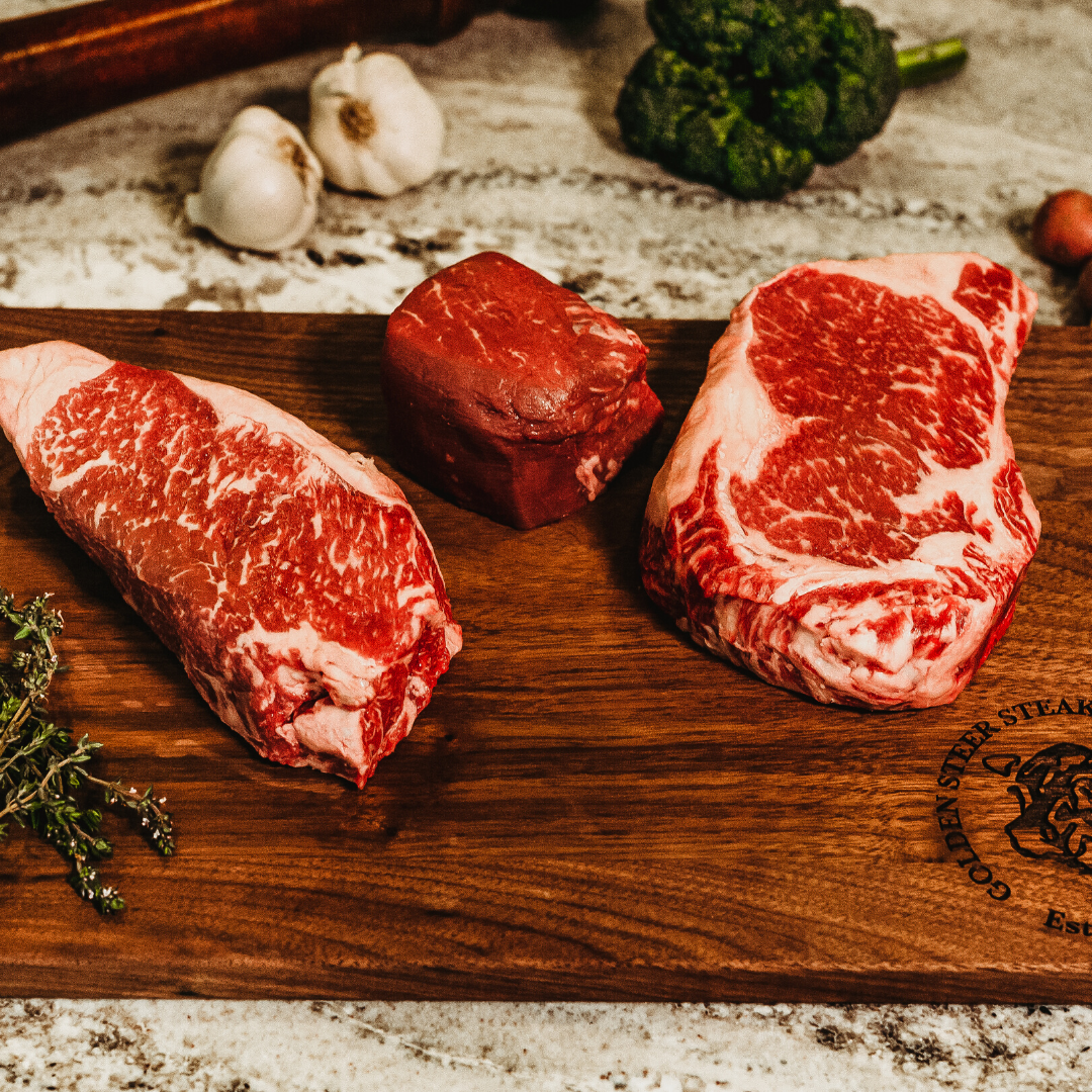 Raw filet, new york, and ribeye on a cutting board surrounded by vegetables