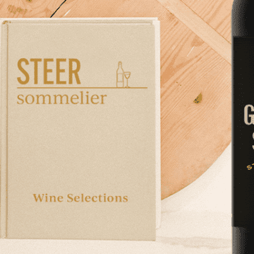Beige book titled 'STEER sommelier' with subtitle 'Wine Selections' beside a partial wine bottle and wooden board.