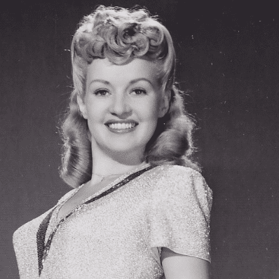 Betty Grable smiling in a 1940s studio portrait with curled hair wearing a sparkly short-sleeve dress.