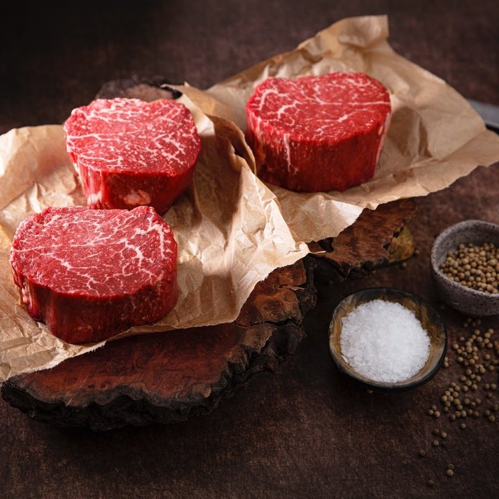 Three marbled beef filet mignon steaks on parchment over a wooden board with a bowl of coarse salt and peppercorns
