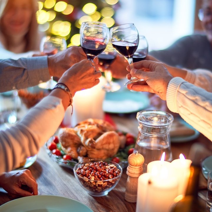 Hands clinking red-wine glasses above a candlelit dinner table with roast chicken, bowls of sides, and soft bokeh lights.