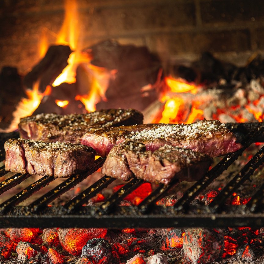 Steaks cooking on a charcoal grill sprinkled with coarse salt, flames and glowing coals visible beneath.