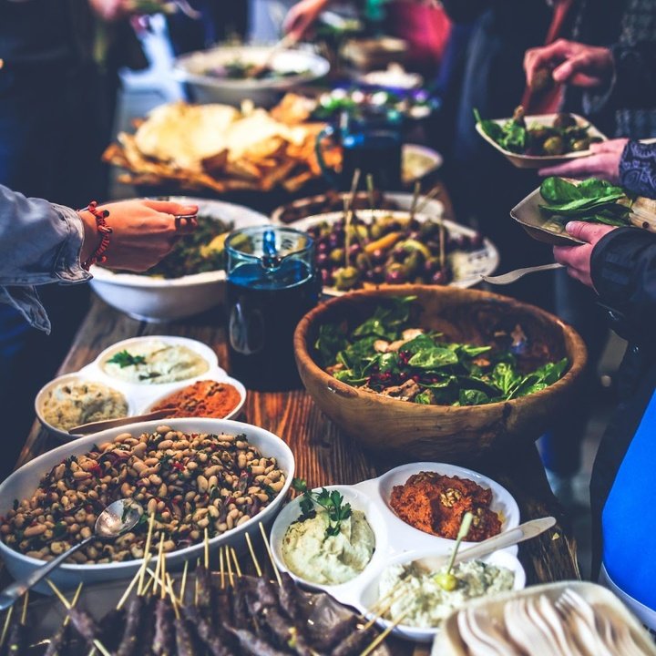 Buffet-style spread on a wooden table: bowls of salad, beans, dips, skewers and people serving themselves.