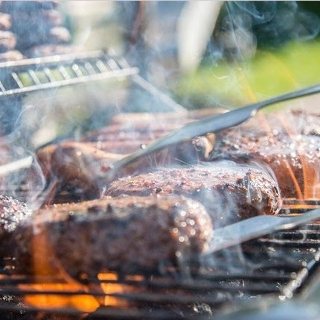 Smoky burger patties on an outdoor grill being flipped with tongs, flames visible under the grate.