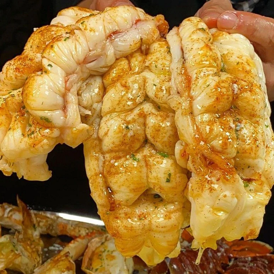 Two cooked lobster tails held by hand, brushed with melted butter and seasoning, juices dripping