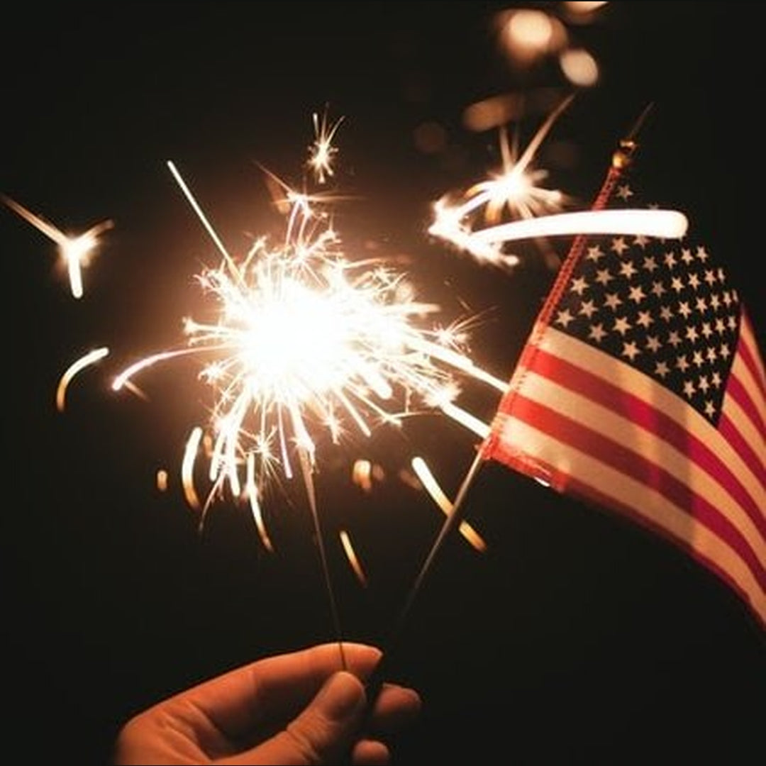 Hand holding two sparklers next to a small American flag glowing against a dark background