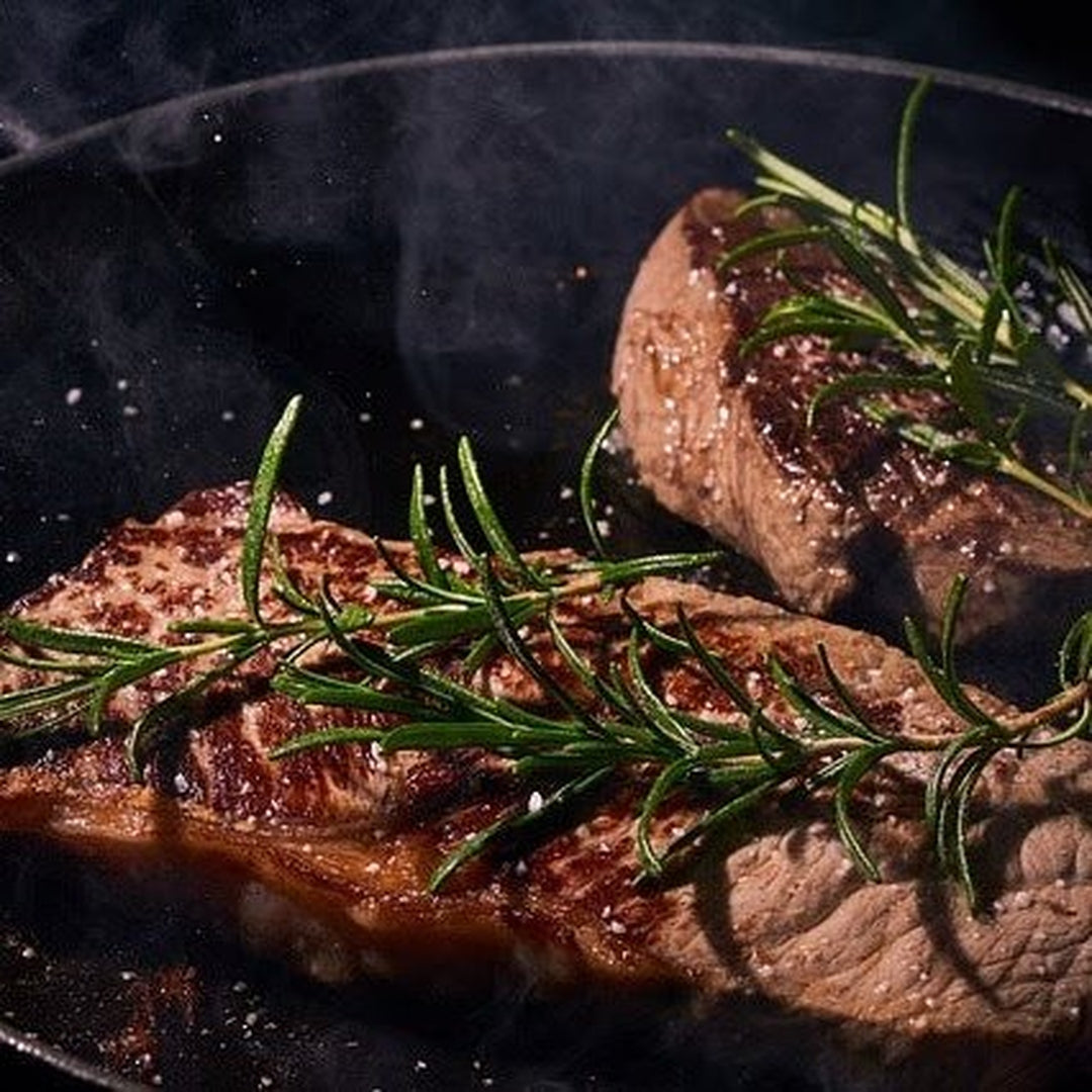 Pan-seared New York steaks in a hot skillet with garlic butter glaze and rosemary sprigs