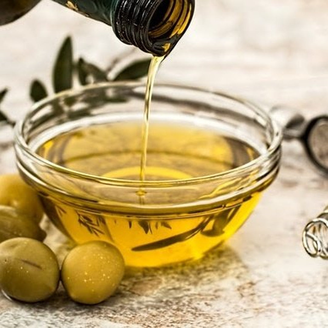 Olive oil being poured into a glass bowl with green olives beside it
