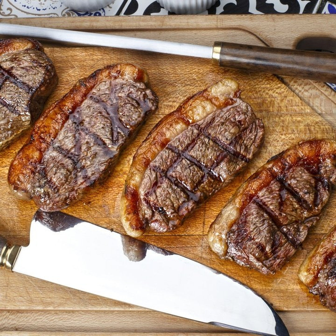 Four grilled ribeye steaks with crosshatch grill marks on a wooden cutting board next to a carving knife