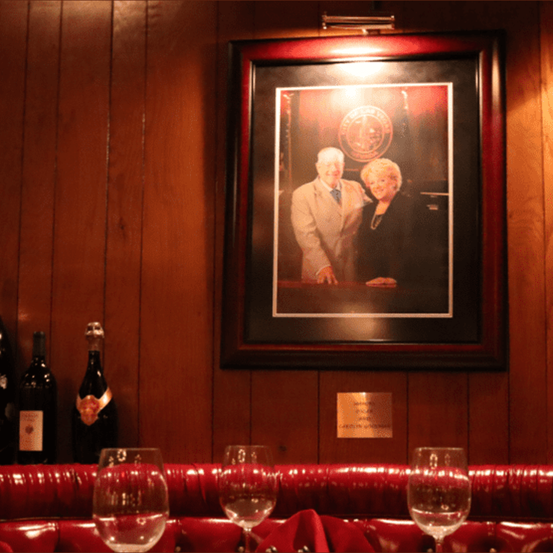 Framed portrait of Oscar and Carolyn Goodman and plaque on wood-paneled wall above a red leather banquette, with wine glasses and bottles in foreground