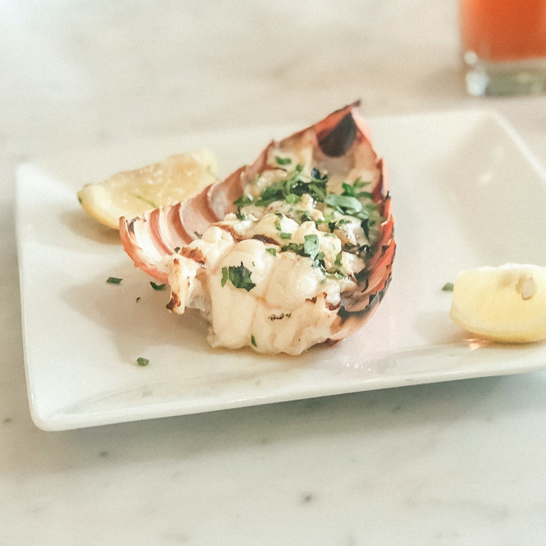 Grilled lobster tail in shell, garnished with chopped parsley and lemon wedges on a white rectangular plate.