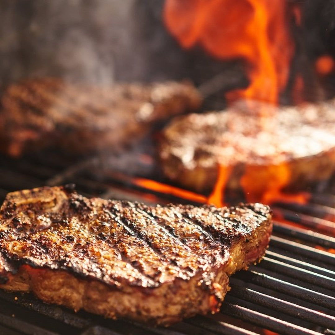 Ribeye steaks searing on a charcoal grill with pronounced grill marks and flames
