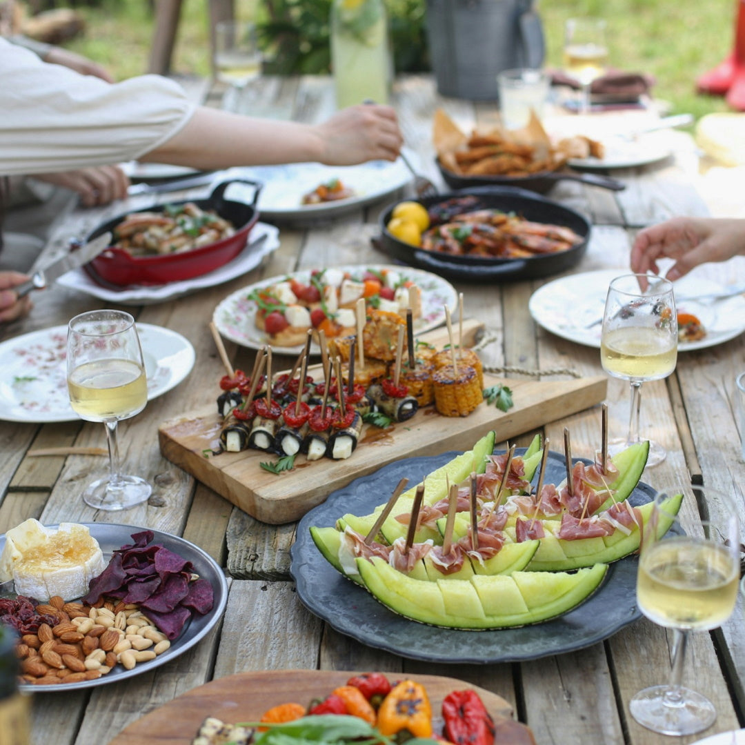 Outdoor wooden table with melon and prosciutto skewers, grilled corn, crostini, nuts and wine glasses.