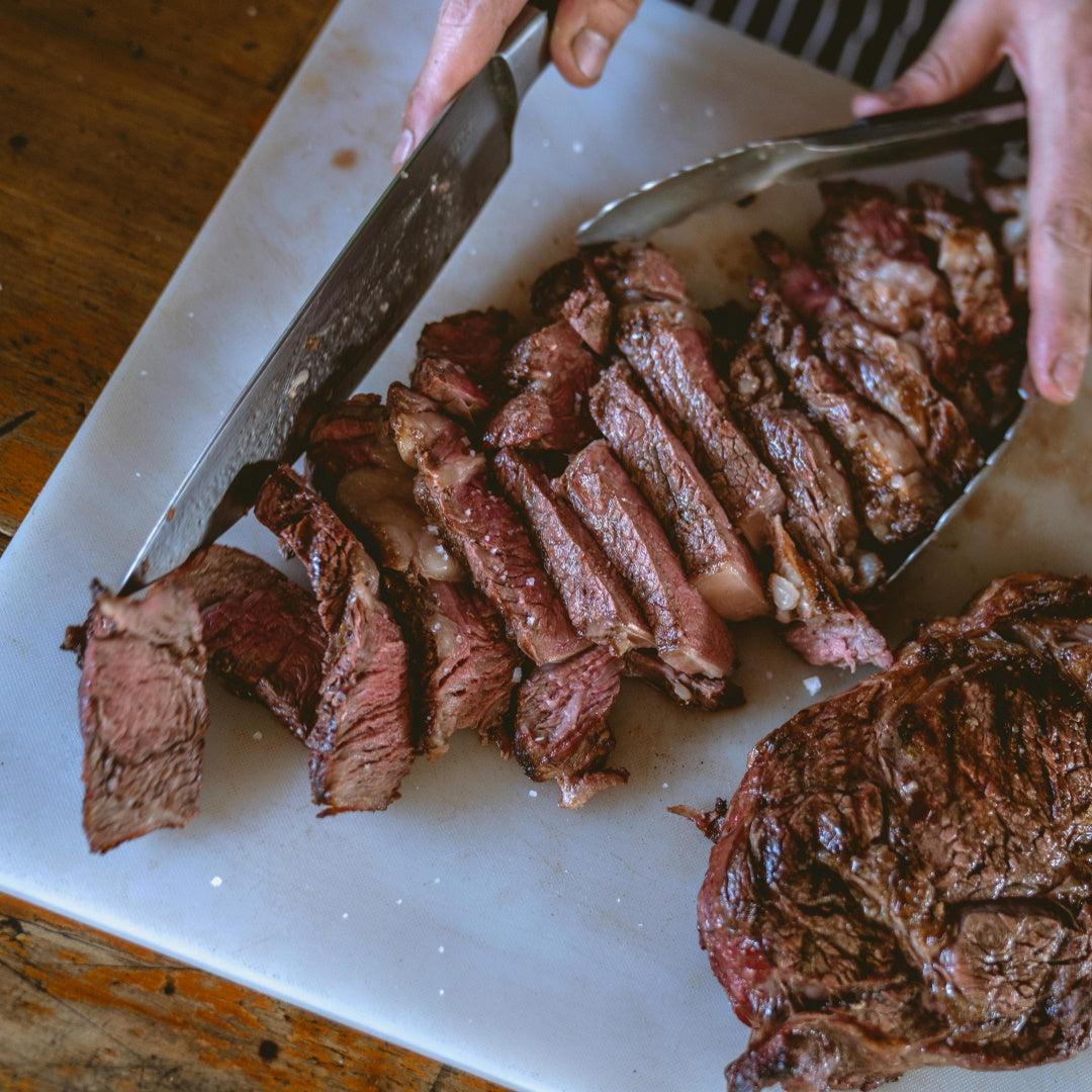 Sliced medium-rare steak being carved with a large knife and tongs on a white cutting board with salt flakes.