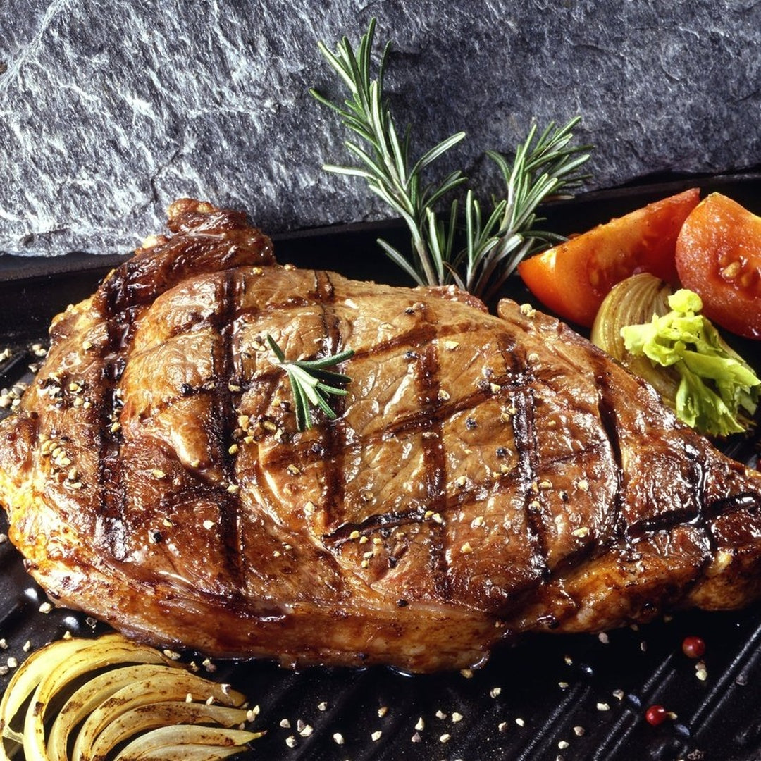 Grilled ribeye steak with crosshatch char marks, rosemary sprigs, tomato wedges and roasted onion on a black grill pan.