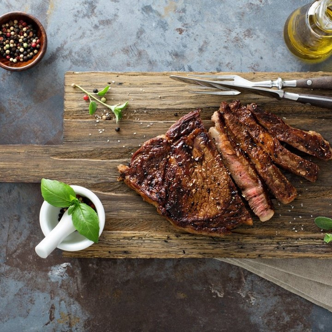 Sliced grilled ribeye on a wooden board with basil in a mortar, peppercorns, a carving fork, and olive oil.