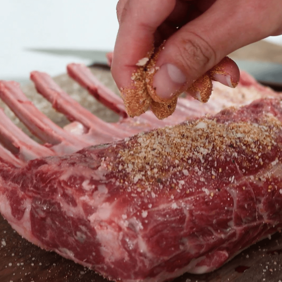 Hand sprinkling seasoning onto a raw rack of lamb, showing exposed rib bones and coarse spice rub on the meat.