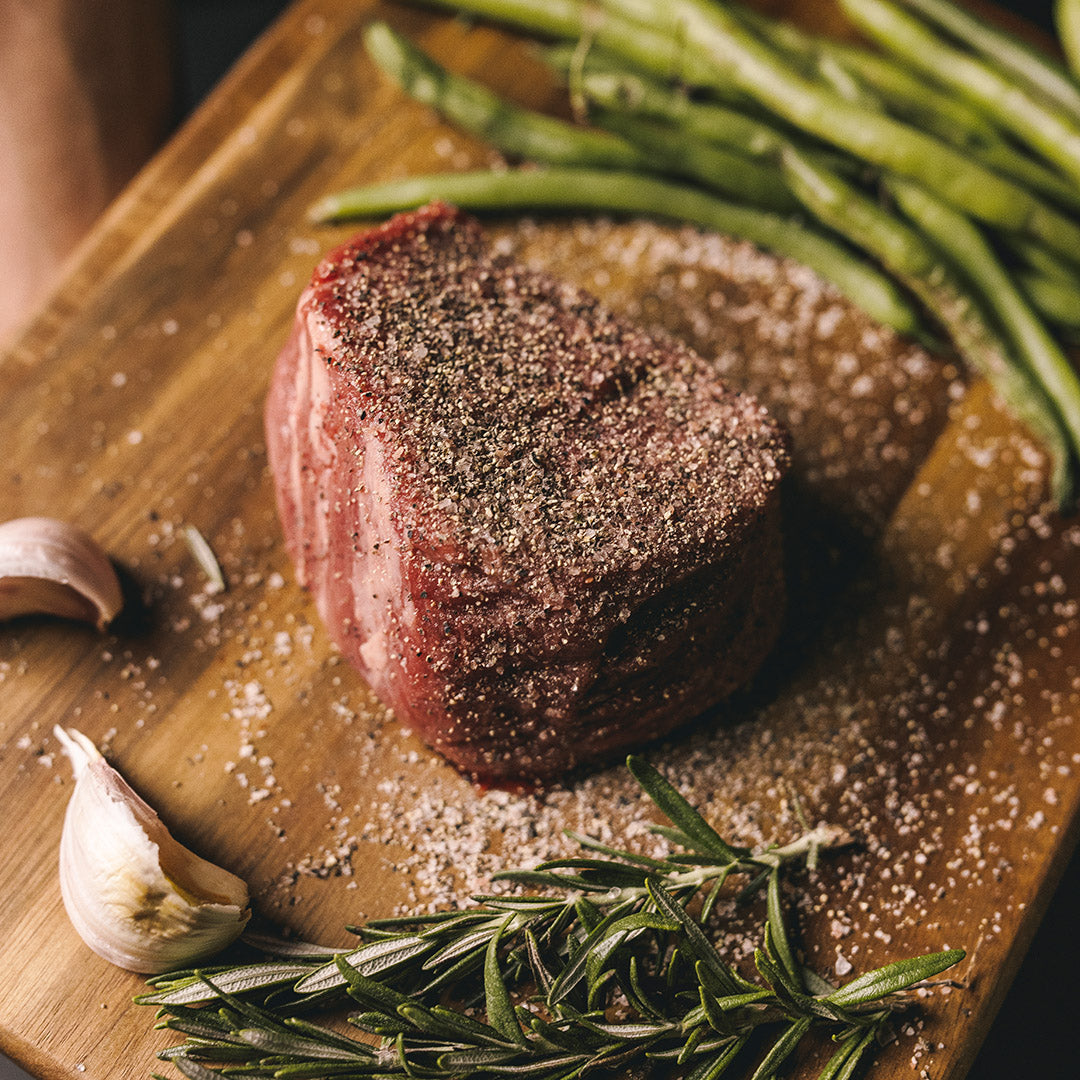 Raw seasoned filet mignon on a wooden board with coarse salt, cracked black pepper, garlic cloves, rosemary and green beans