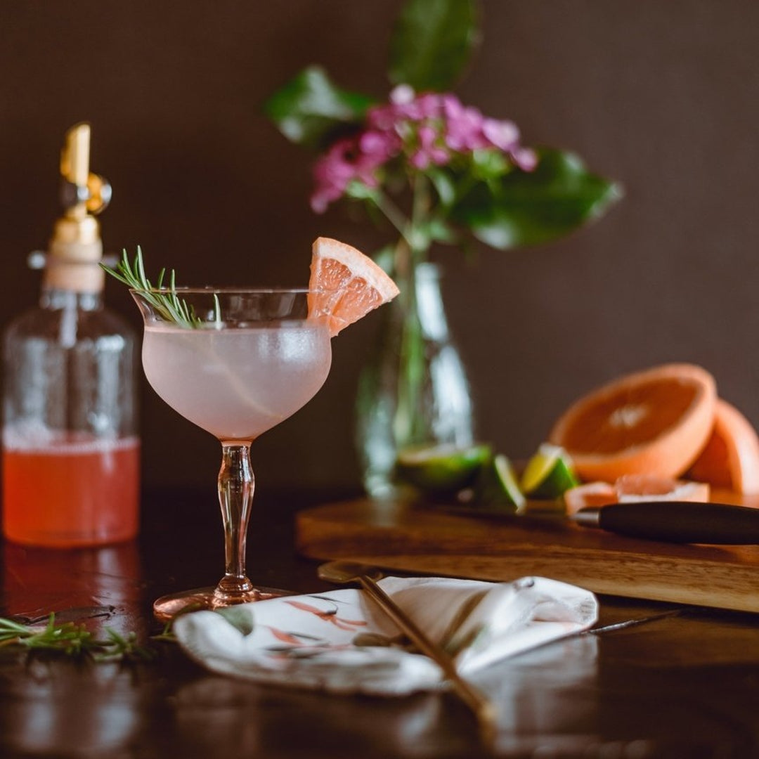 Pale pink grapefruit cocktail in a coupe glass garnished with rosemary and a grapefruit wedge, with cutting board and halved fruit.