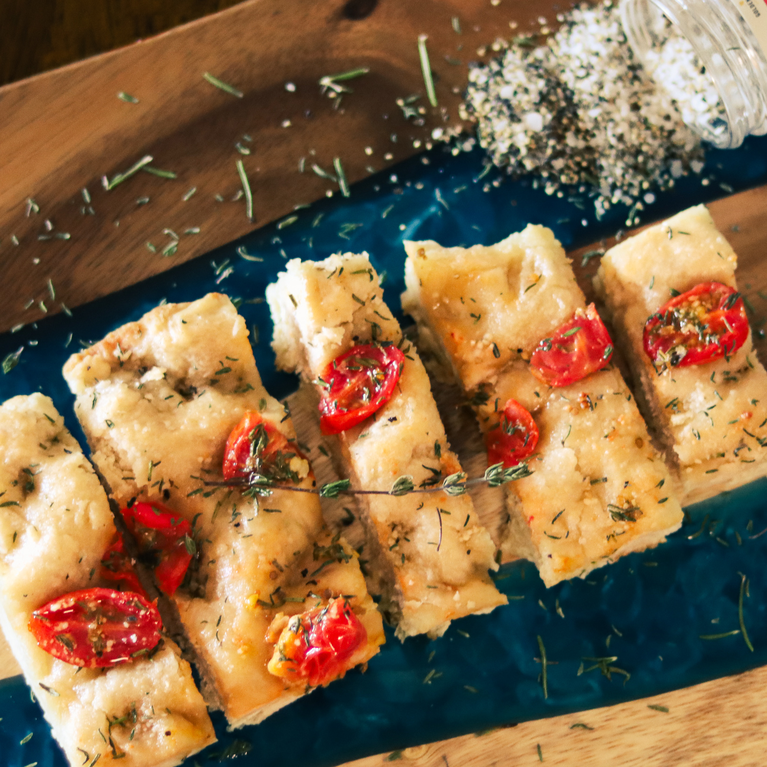 Sliced focaccia with cherry tomato halves, herbs and coarse salt on a blue serving board.