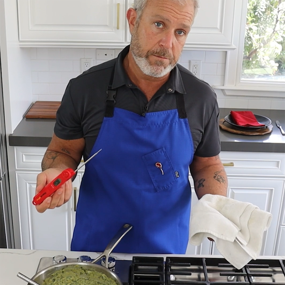 Person in blue apron holding a red digital probe thermometer over a pot on the stove, towel in other hand.