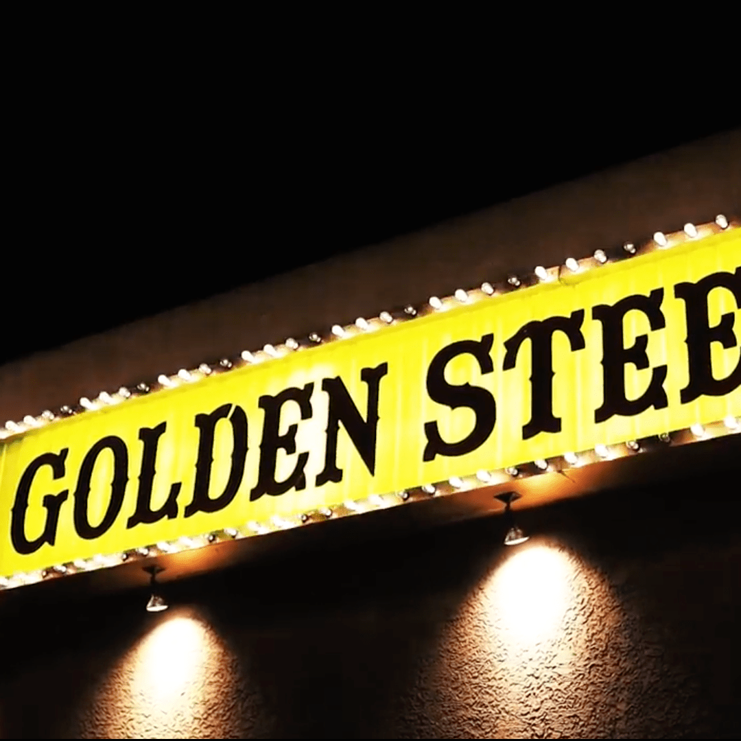 Yellow marquee sign reading 'GOLDEN STEE' in large black letters, bordered by bulbs with two wall spotlights below.