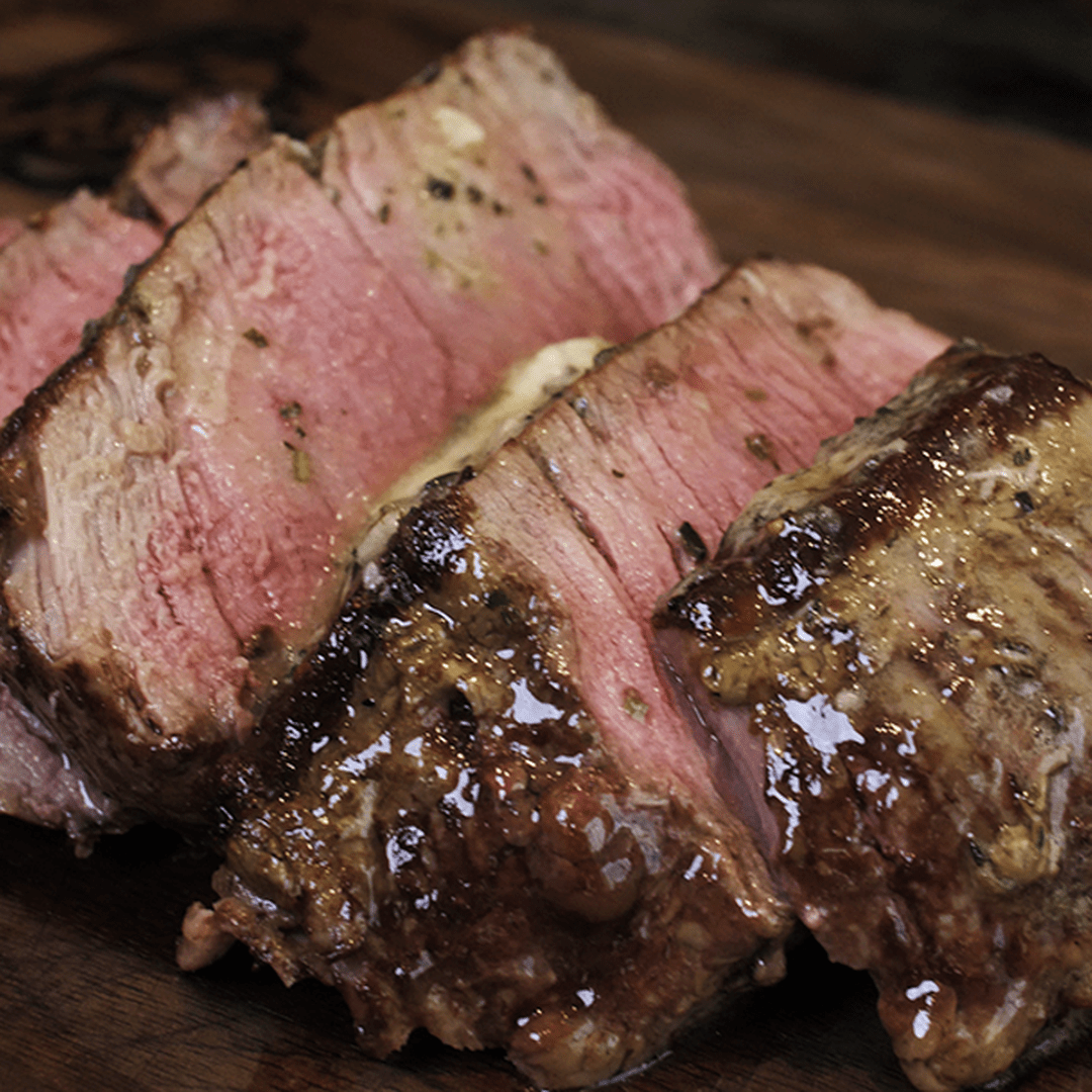 Sliced medium-rare beef roast with melting herb butter and juices on a wooden cutting board