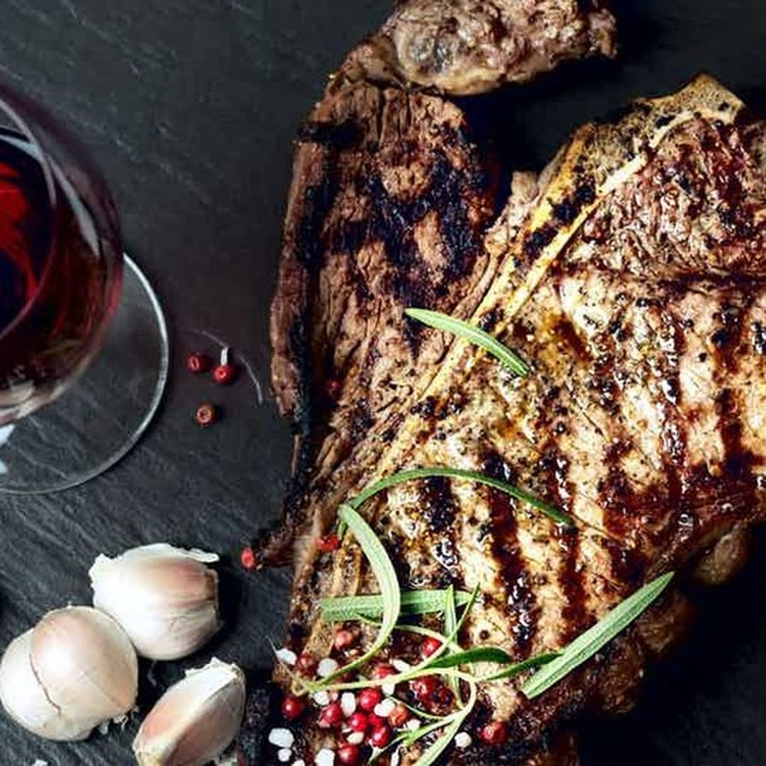 Grilled bone-in steak with grill marks, rosemary sprigs, garlic cloves, pink peppercorns and a glass of red wine on a slate