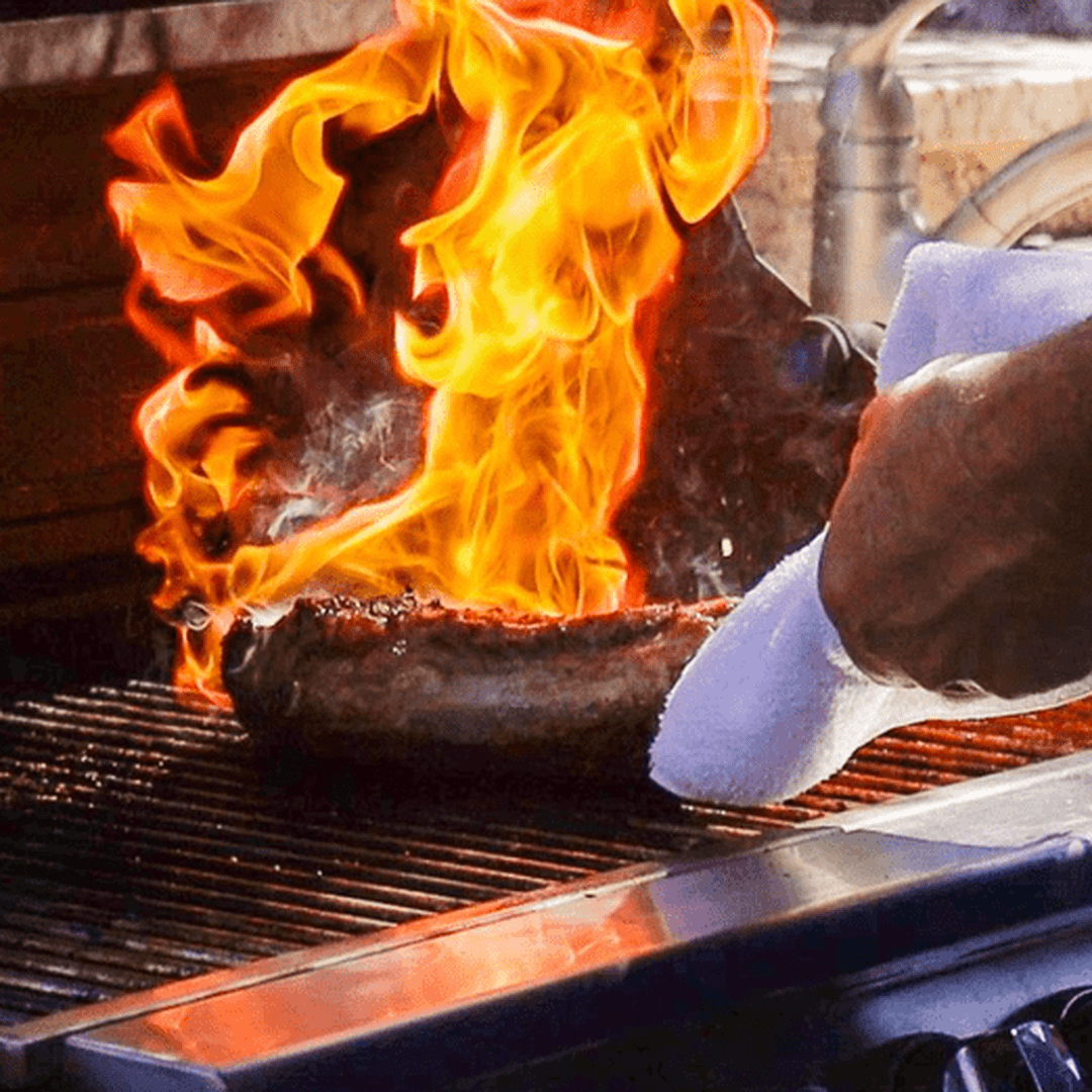 Meat sizzling on a grill with tall orange flames and a hand holding a cloth at the grill edge.