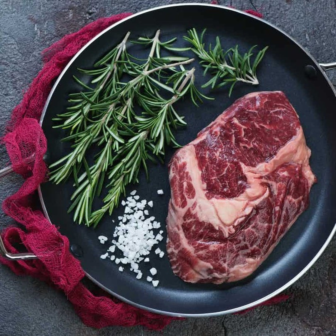 Ribeye steak in a skillet with rosemary sprigs and coarse salt on a red cloth.
