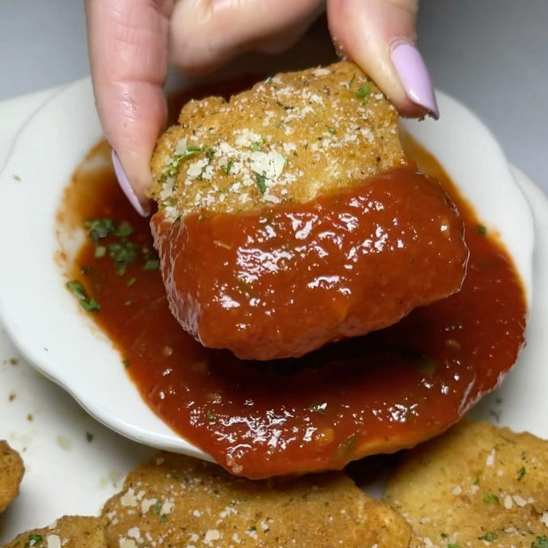 Breaded toasted ravioli being dipped into marinara sauce on a small white dish, garnished with grated cheese and parsley.