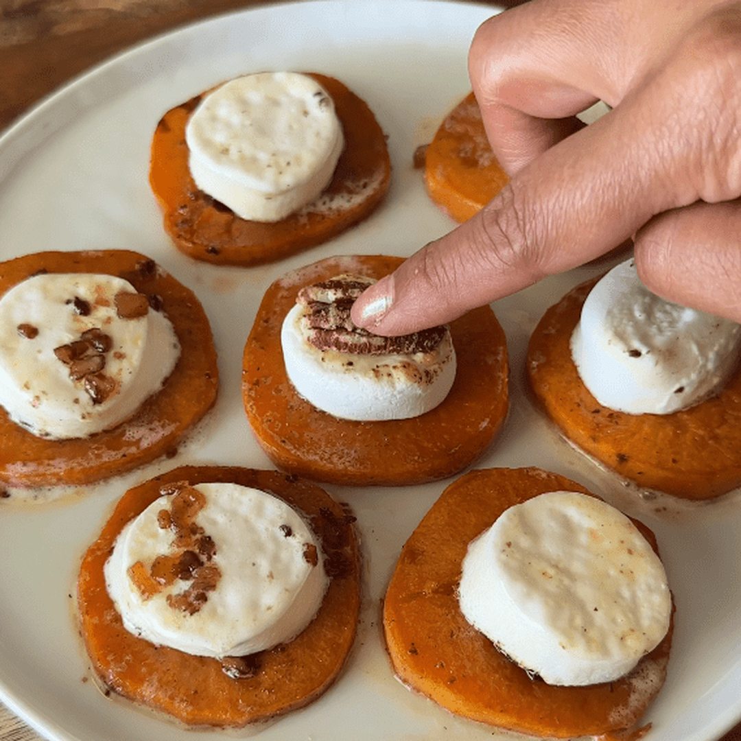 Roasted sweet potato rounds topped with marshmallows and chopped pecans, one marshmallow being pressed by a finger.