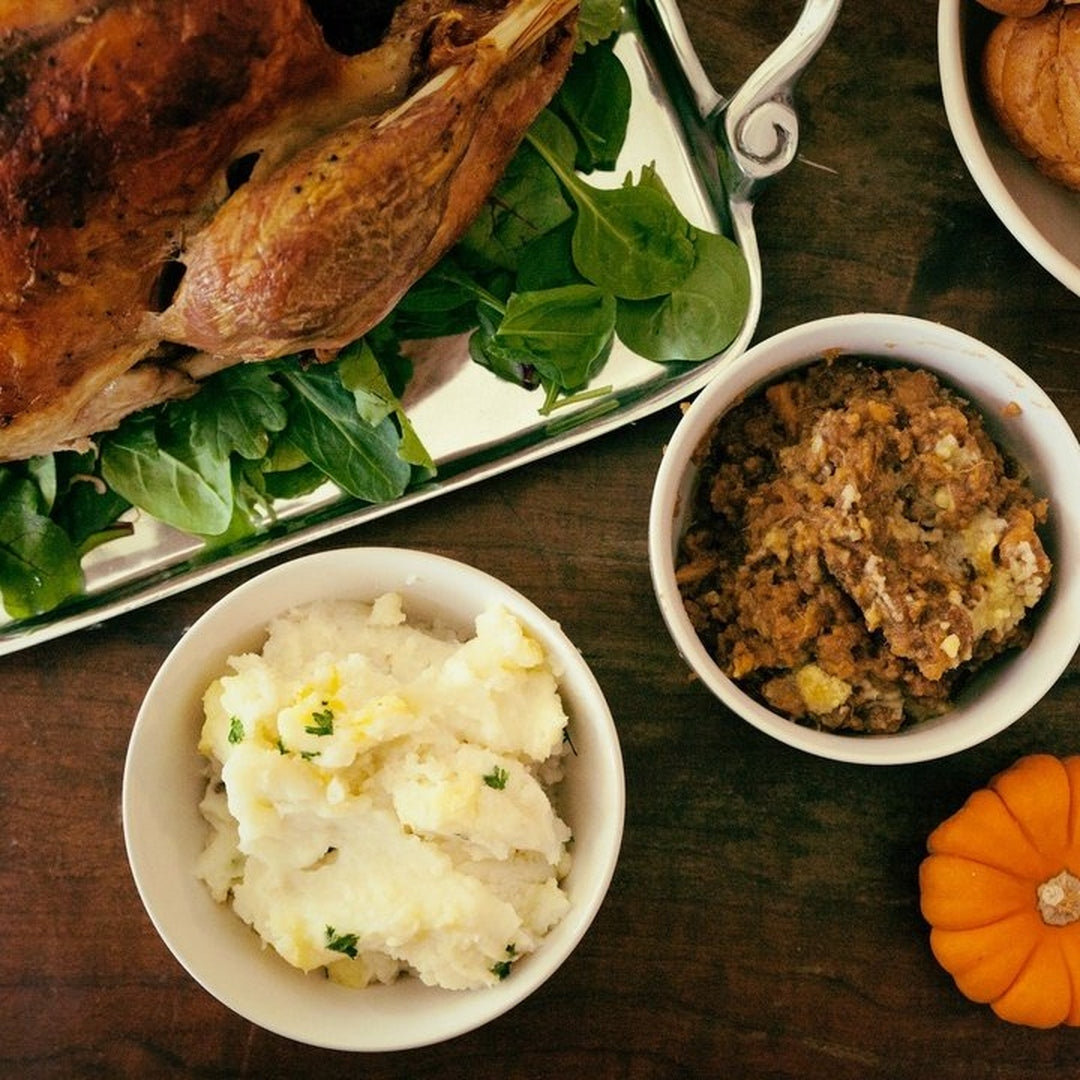 Overhead Thanksgiving spread: roasted turkey on greens, bowl of mashed potatoes with butter, bowl of stuffing, small pumpkin.