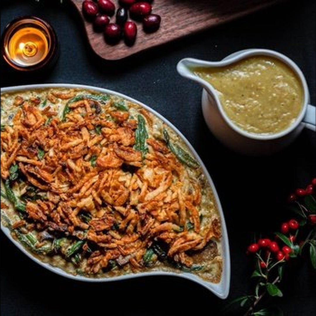Oval green bean casserole topped with crispy fried onions, white gravy pitcher, red cranberries and a lit candle on dark table.