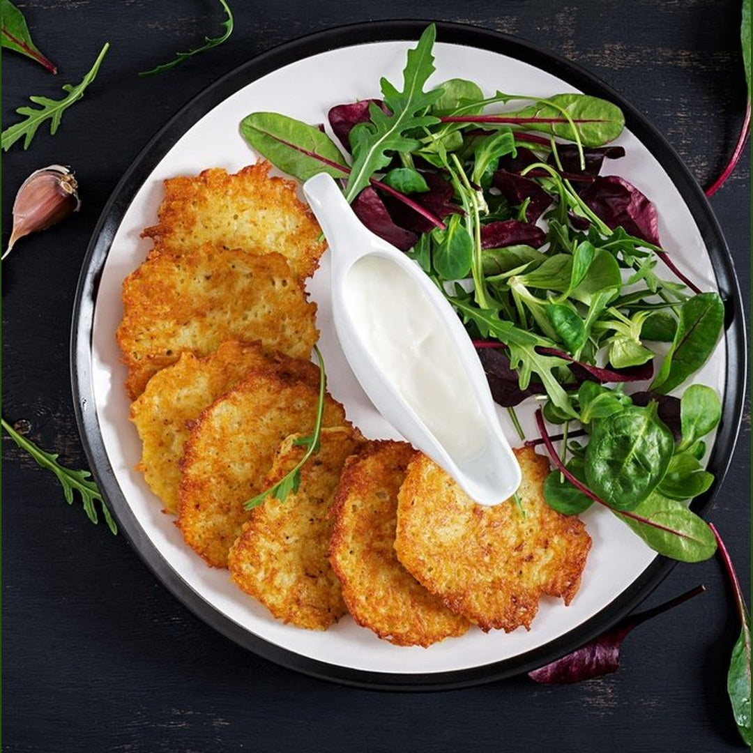 Stack of crispy golden potato latkes on a white plate with sour cream in a gravy boat and mixed salad greens.