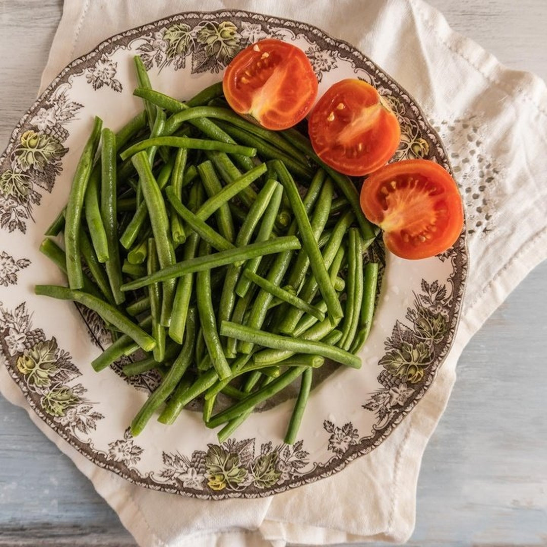 Steamed green beans piled on a patterned china plate with three halved tomatoes on a cloth napkin.
