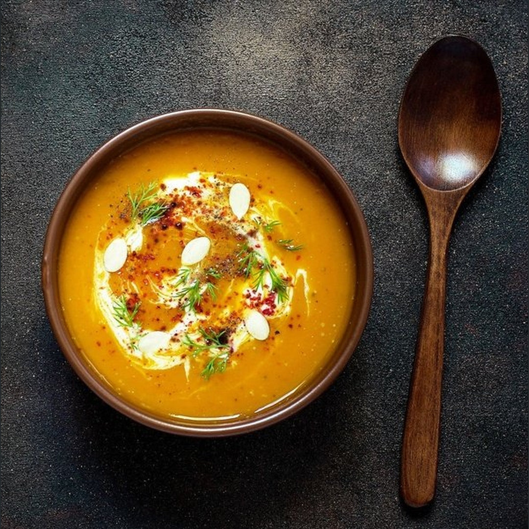 Bowl of roasted butternut squash soup garnished with cream swirl, pumpkin seeds, herbs and red pepper flakes; wooden spoon at right.