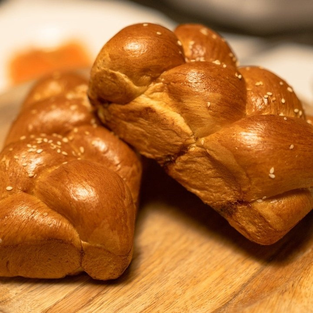 Two golden braided challah loaves with sesame seeds on a wooden cutting board