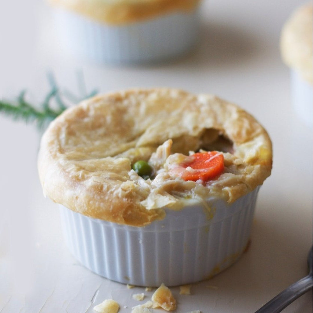 Mini chicken pot pie in white ramekin with torn golden crust, visible peas and carrot slice, flaky crumbs on table
