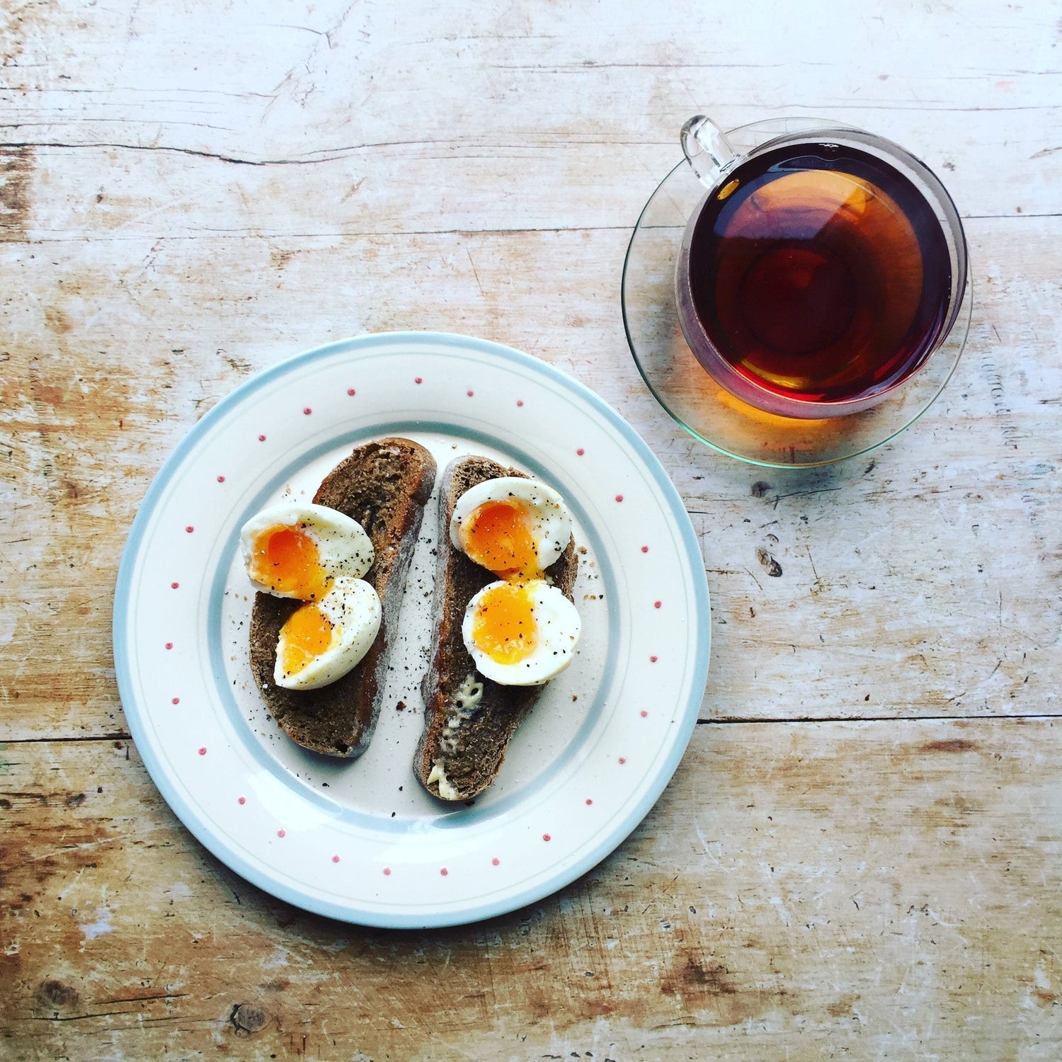 Two slices of toast topped with halved soft‑boiled eggs on a white plate; glass cup of tea on a wooden table.