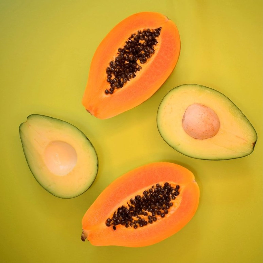 Overhead view of two halved papayas with black seeds and two halved avocados, one with pit, on a yellow-green background