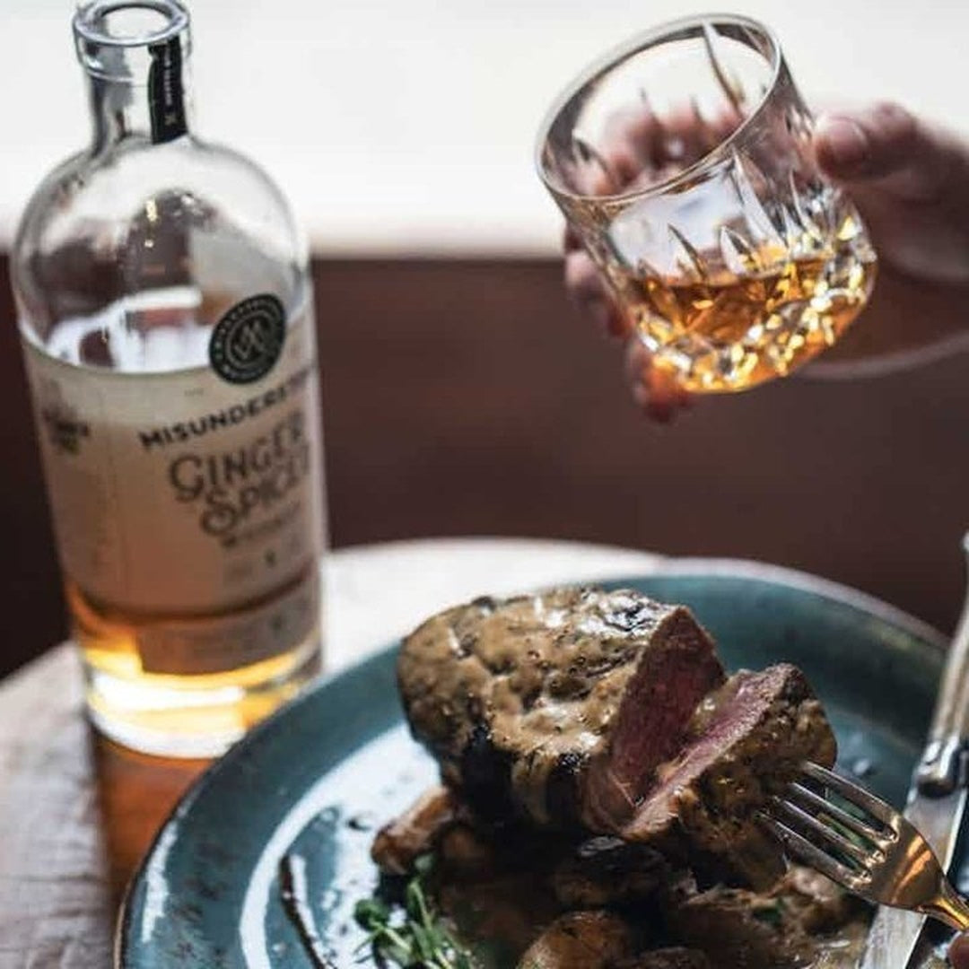 Steak on a plate with a bottle labeled 'MISUNDERSTOOD GINGER SPICE' and a hand holding a whiskey glass