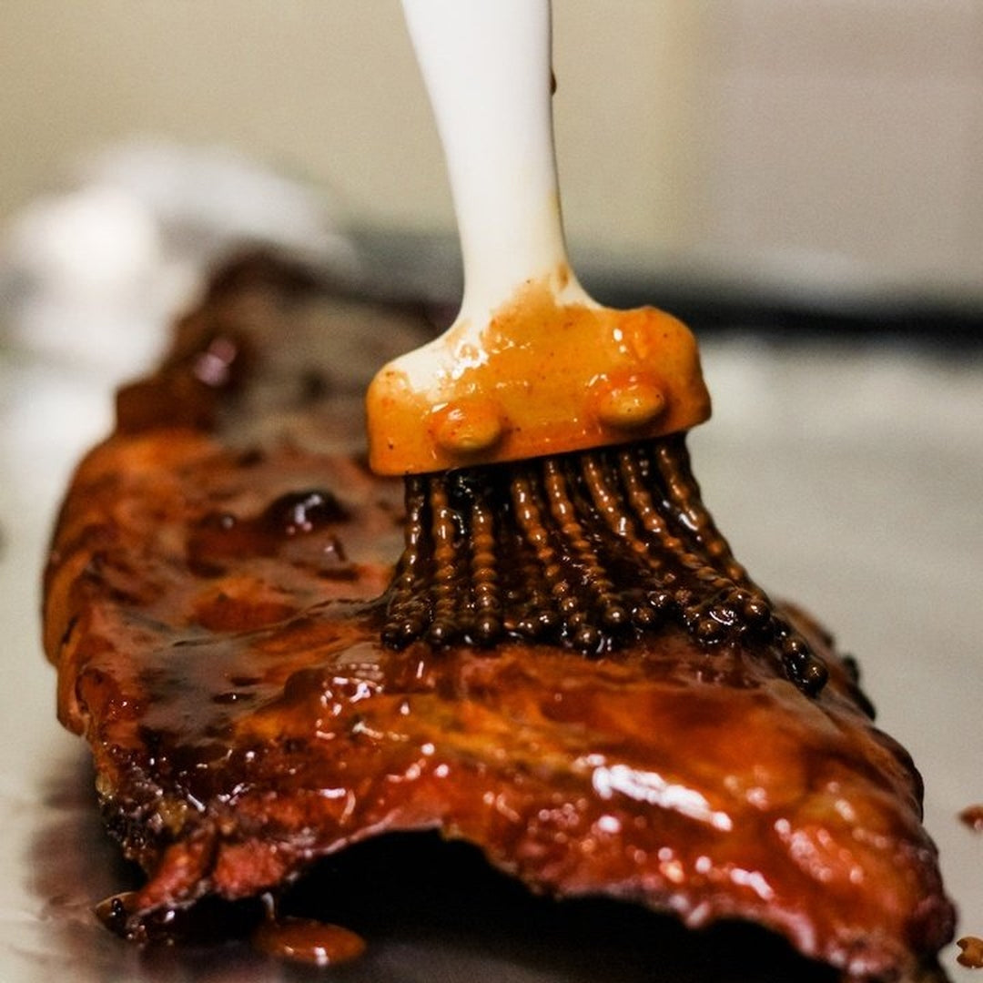 Basting brush spreading glossy barbecue sauce over a rack of smoked pork ribs, close-up of sauce-coated bristles and glazed meat